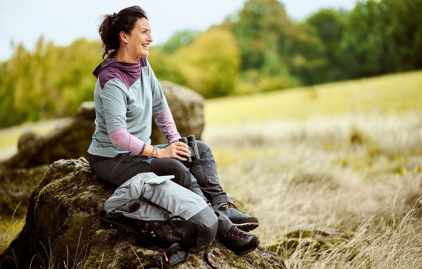 Wanderin sitzt auf einem Stein mit Fernglas in der Hand glücklich in die Ferne blickend