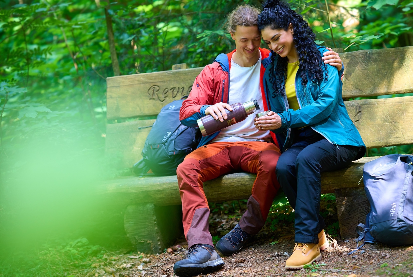 Glückliches Paar im Wanderoutfit sitzend auf einer Bank im Wald bei einer Rast. Mann schenkt frischen Tee aus