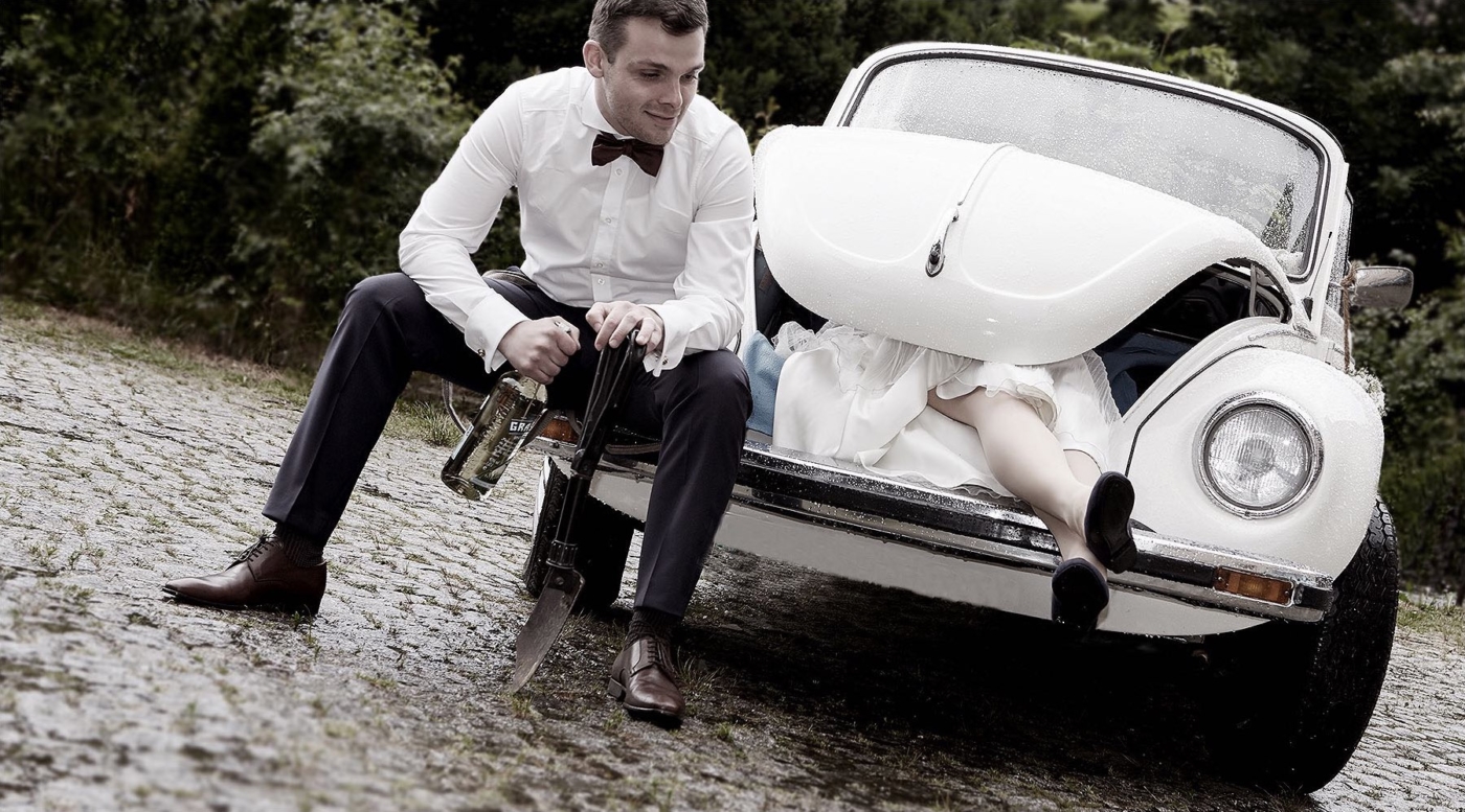 Die Braut liegt im Kofferraum eines VW Käfers und der Bräutigam daneben mit Whiskyflasche und Spaten n der Hand