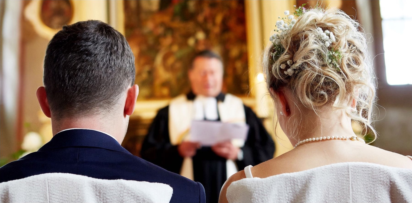 Rückenportrait eines Brautpaars. Im Hintergrund der Pfarrer unscharf während der Trauzeremonie.