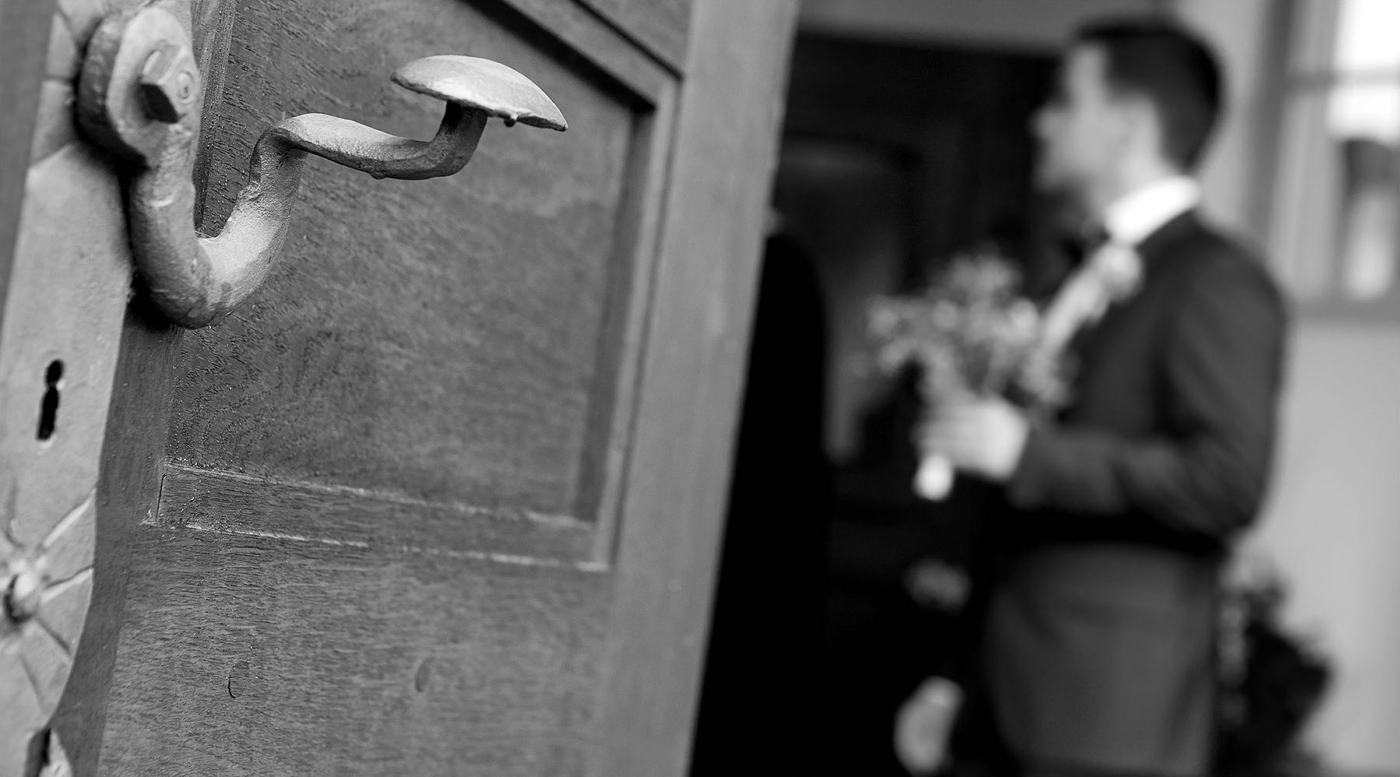 Schwarz Weiss Aufnahme der Türklinke einer Kirche im Hintergrund unscharf der Bräutigam