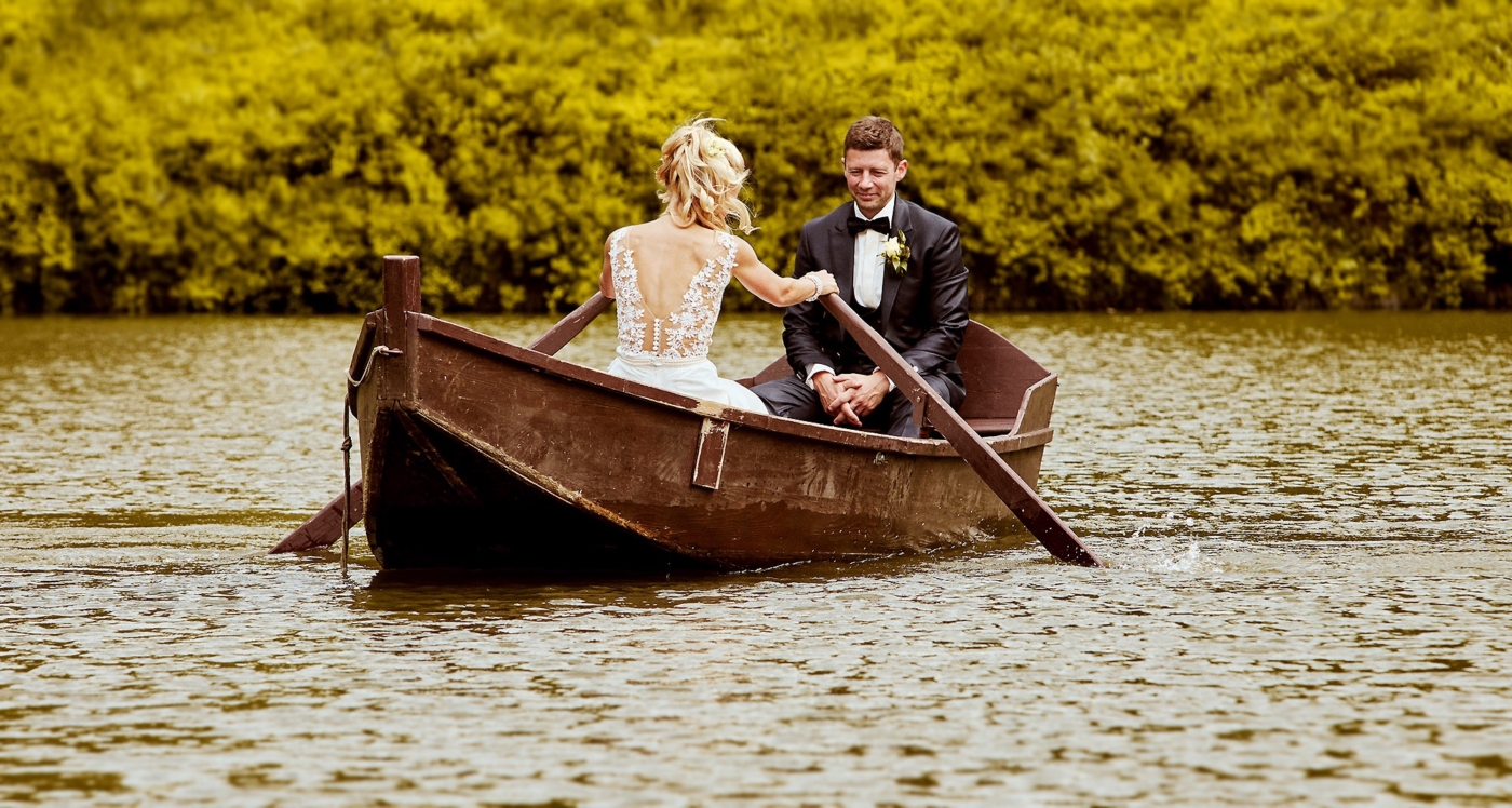 Die Braut rudert ein altes Boot auf einem romantischen See und der Bräutigam sitzt ihr gegenüber in nachdenklicher Pose
