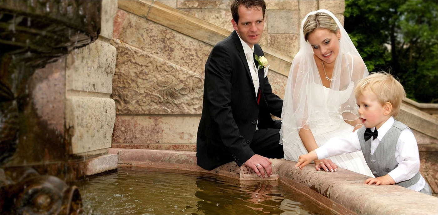 Ein junges Hochzeitspaar sitzt mit Ihrem Kind an einem Brunnen im Park während das Kind auf das Wasser zeigt