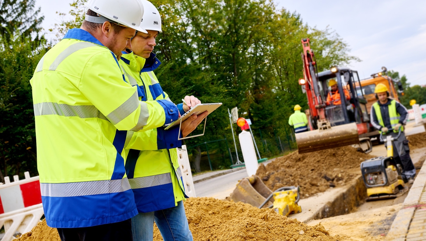 2 Männer mit Helm an einer Strassen Baustelle im prüfen der Bauunterlagen. Im Hintergrund Mann mit Verdichter