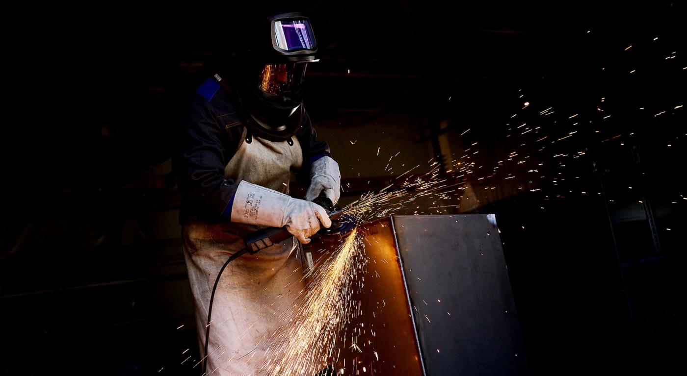 Mann mit Arbeitshandschuhen und Schweisserhelm beim bearbeiten einer Metallbox mit Trennschleifer