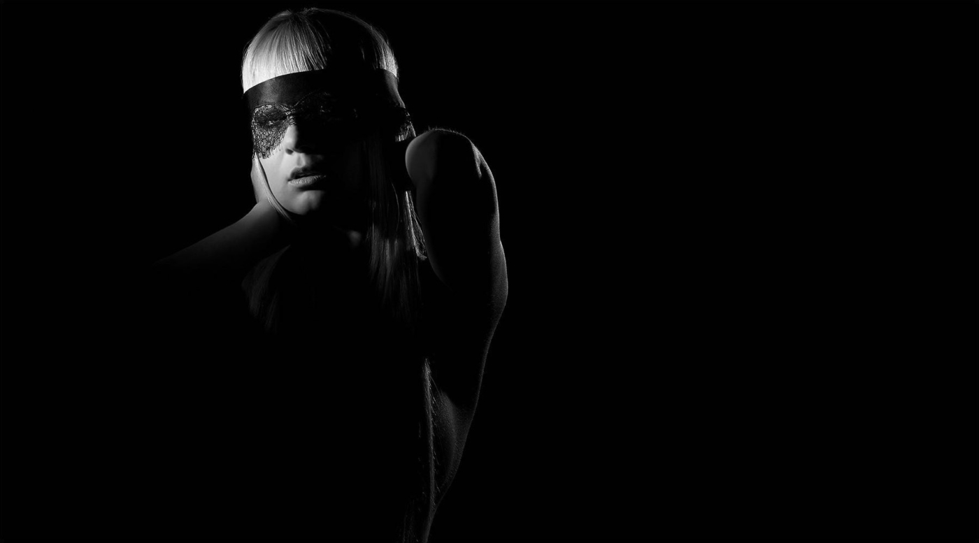 Portrait einer blonden Frau mit gestickter Maske vor den Augen und Händen am Kopf